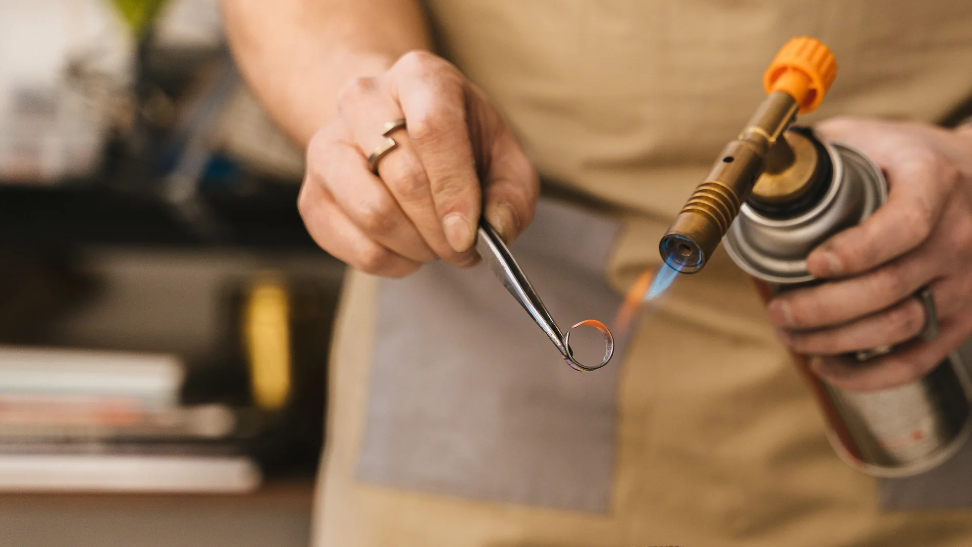 A silversmith holding a small silver ring in tweezers in front of a gas torch flame, at a workbench with a soldering block
