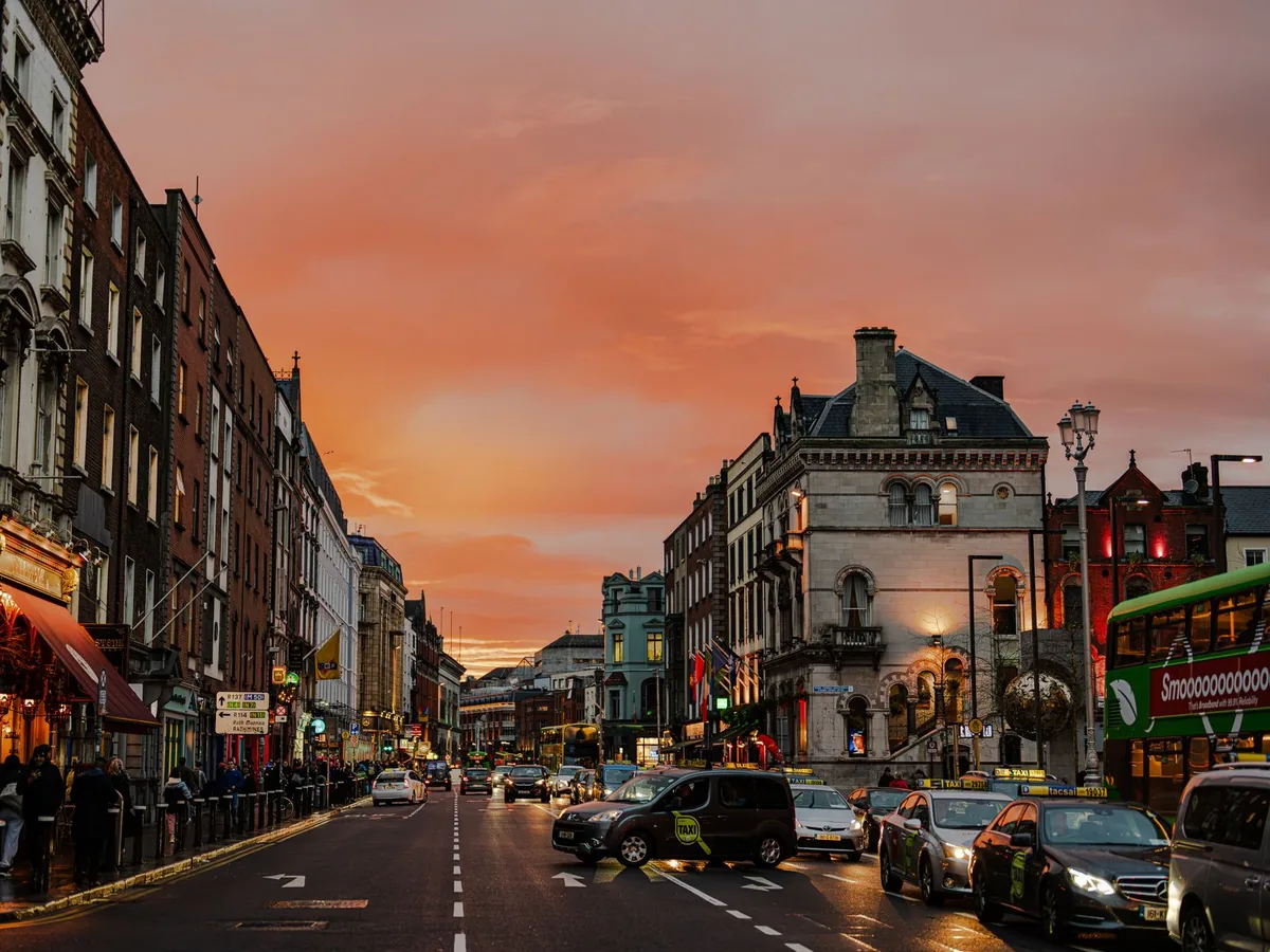 A busy Dublin street at sunset with traffic, a double-decker bus and a lit stone building ahead